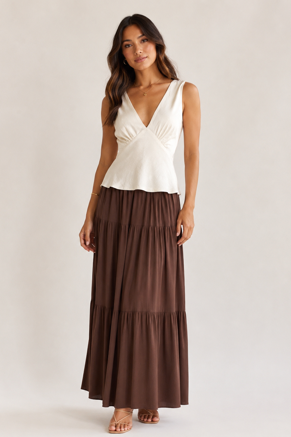 A woman modeling a sleeveless white wrap-style top paired with a long brown tiered maxi skirt, standing against a plain light beige studio background.