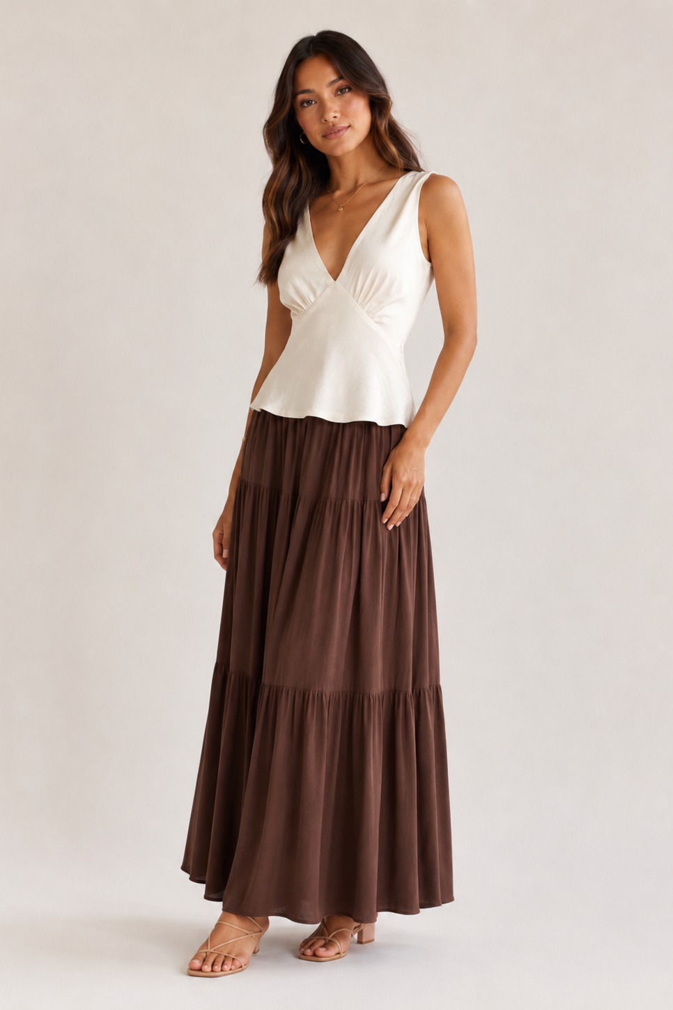 A woman modeling a sleeveless white V-neck top paired with a long brown tiered maxi skirt, standing against a plain light beige studio background.