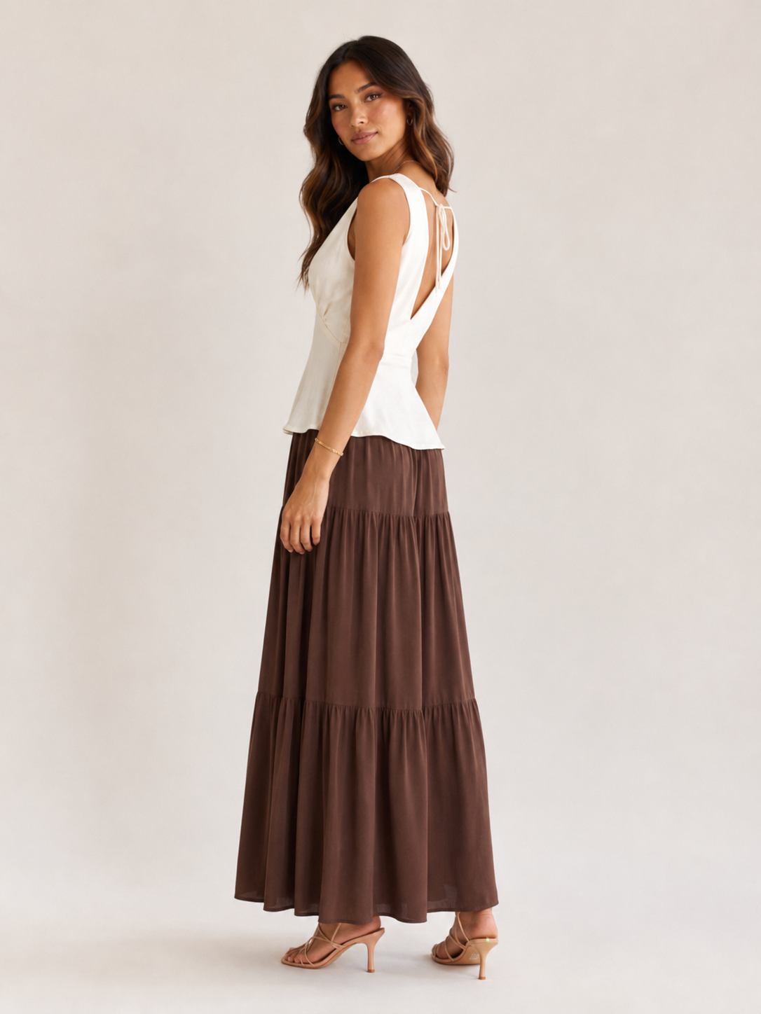 A woman modeling a sleeveless white top with an open back and a long brown tiered maxi skirt, shown from the back against a plain light beige studio background.