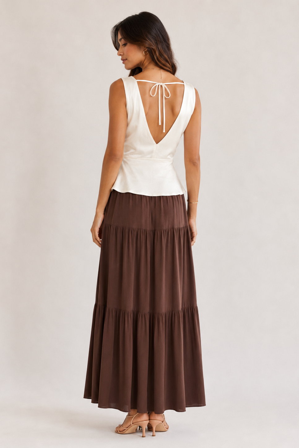 A woman shown from the back wearing a sleeveless white top with a tie-back neckline and a long brown tiered skirt, standing against a plain light studio background.
