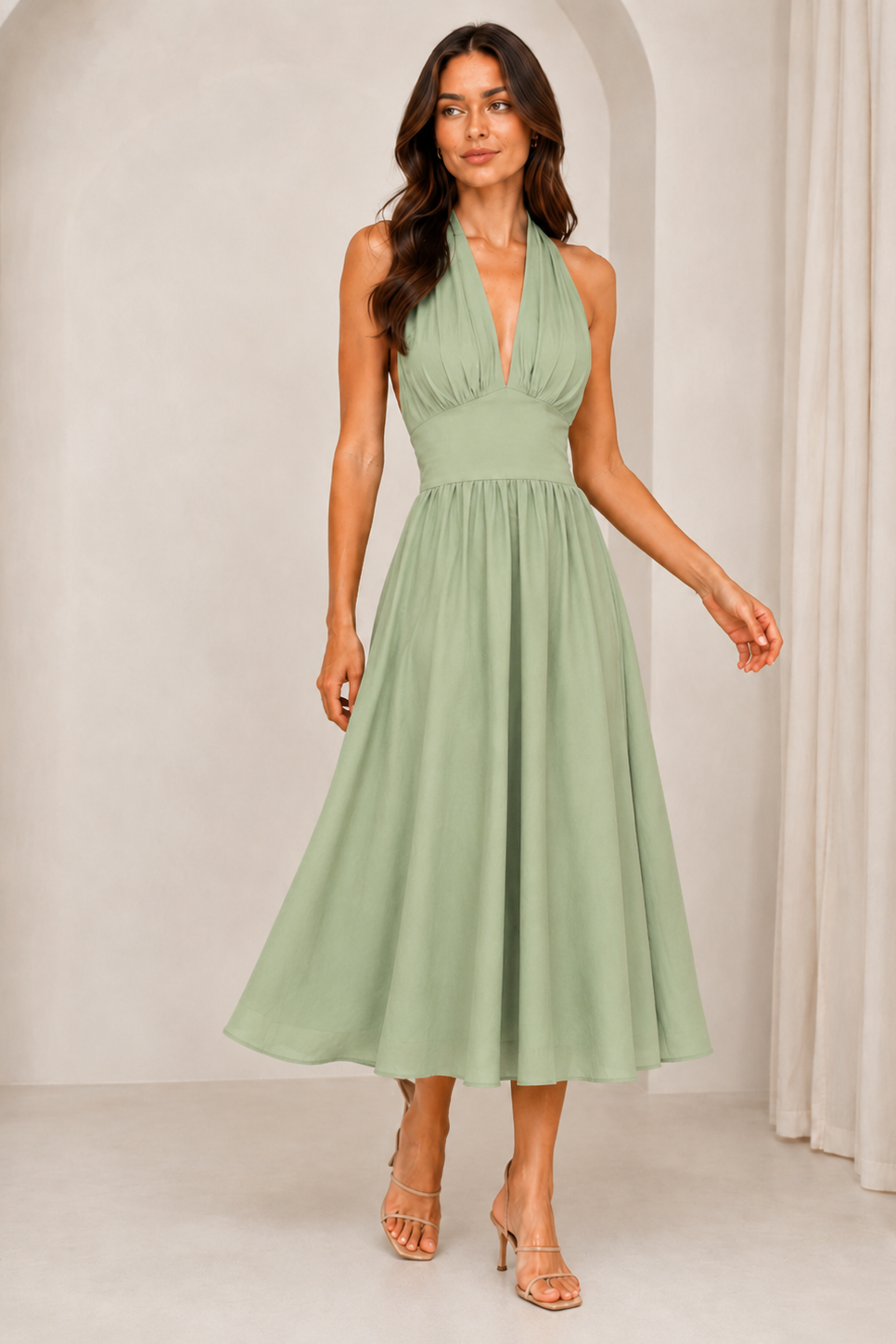 A woman modeling a sage green halter midi dress with a gathered waist and flowing skirt, paired with clear high-heeled sandals in a neutral studio setting.