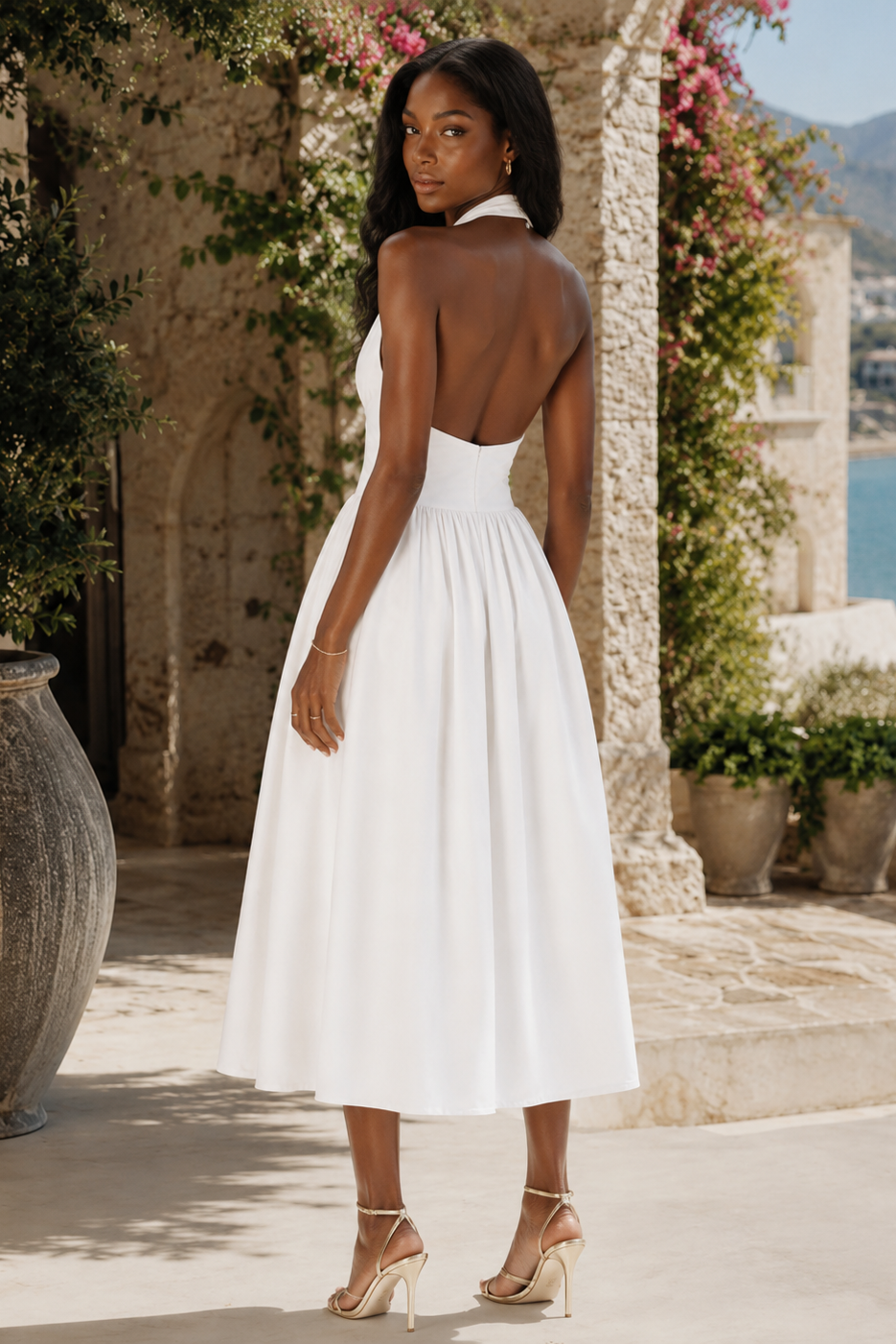 A woman wearing a white sleeveless backless midi dress with a pleated skirt, standing outdoors in front of a stone wall with greenery and a view of the sea.