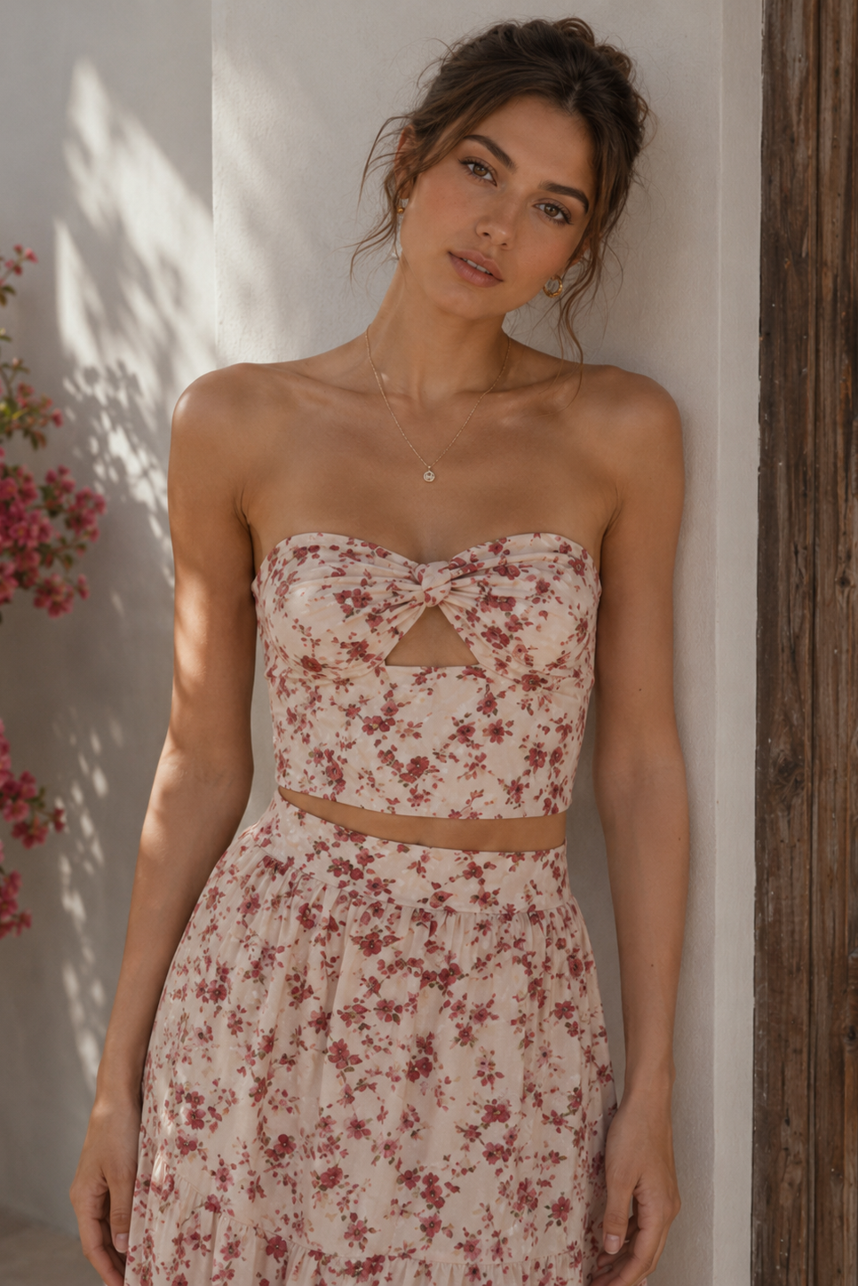 Woman wearing a floral dress standing against a white wall with flowers and a wooden door in the background.
