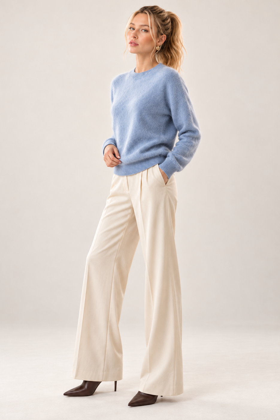 A woman modeling a light blue knit crewneck sweater and cream wide-leg trousers with dark brown pointed-toe heels, standing against a neutral light gray studio background.