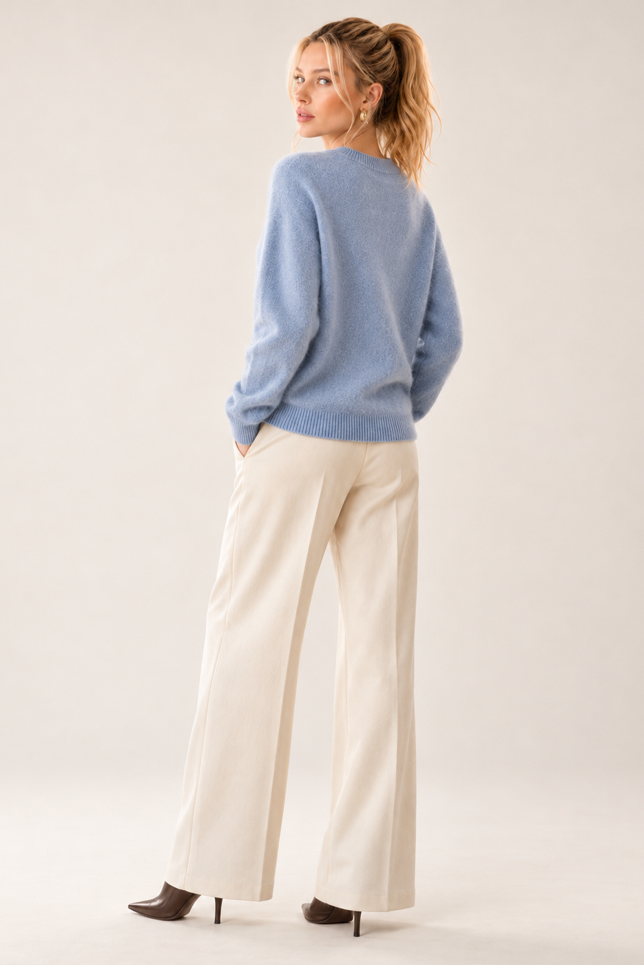 A woman stands in a studio wearing a light blue knit sweater, cream wide-leg trousers, and dark brown pointed heels, photographed from the back against a plain light beige background.