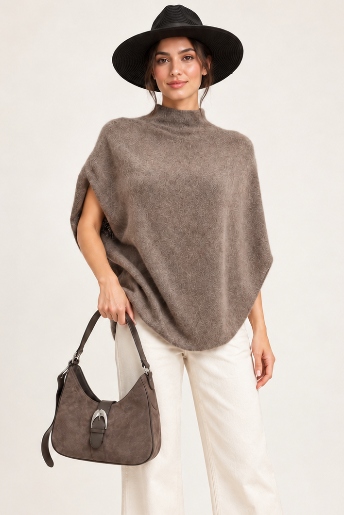 A model wearing a taupe brown fuzzy poncho sweater, white wide-leg pants, brown ankle boots, a black wide-brim hat, and holding a taupe brown handbag against a light beige studio background.