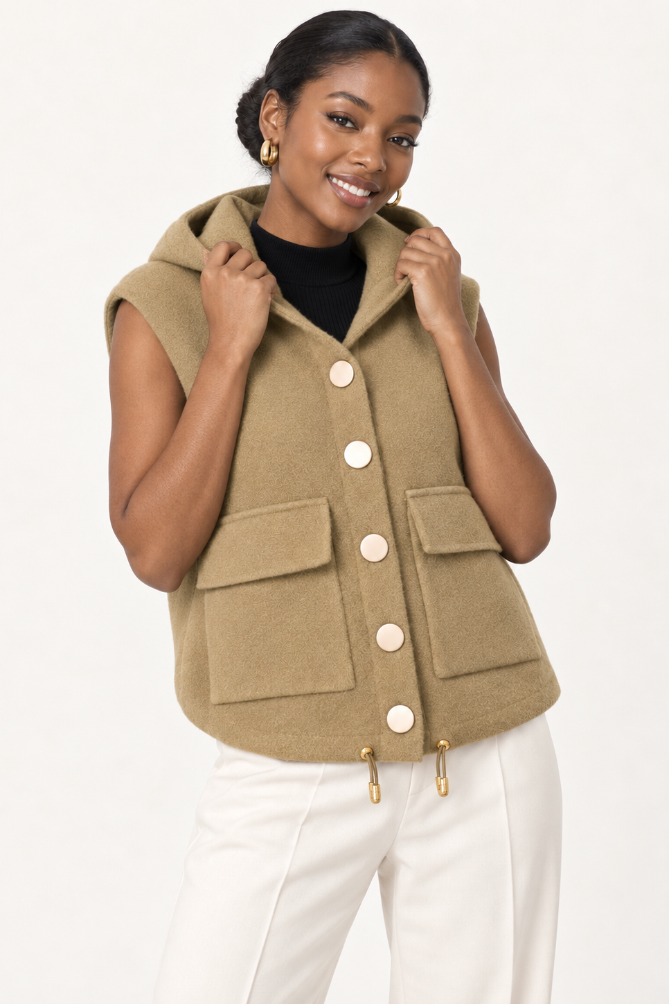 A woman modeling a sleeveless olive green hooded vest with large front buttons, flap pockets, and drawstring details, paired with white pants and a black top against a plain light background.