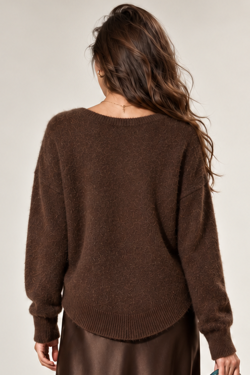 A woman shown from behind wearing a brown knit sweater, a brown satin midi skirt, and brown heeled boots, while holding a brown handbag with a blue patterned scarf.