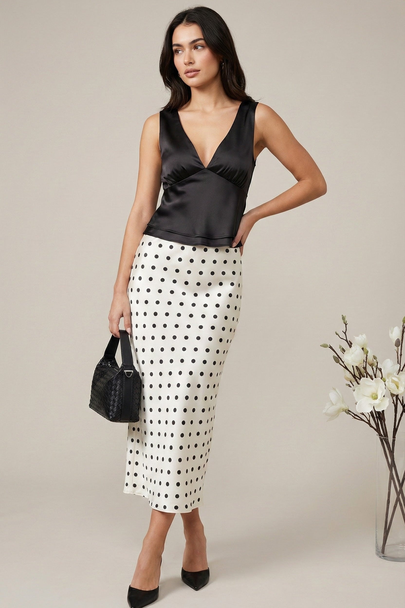 Woman wearing a black satin top and polka dot skirt holding a black handbag against a neutral background.