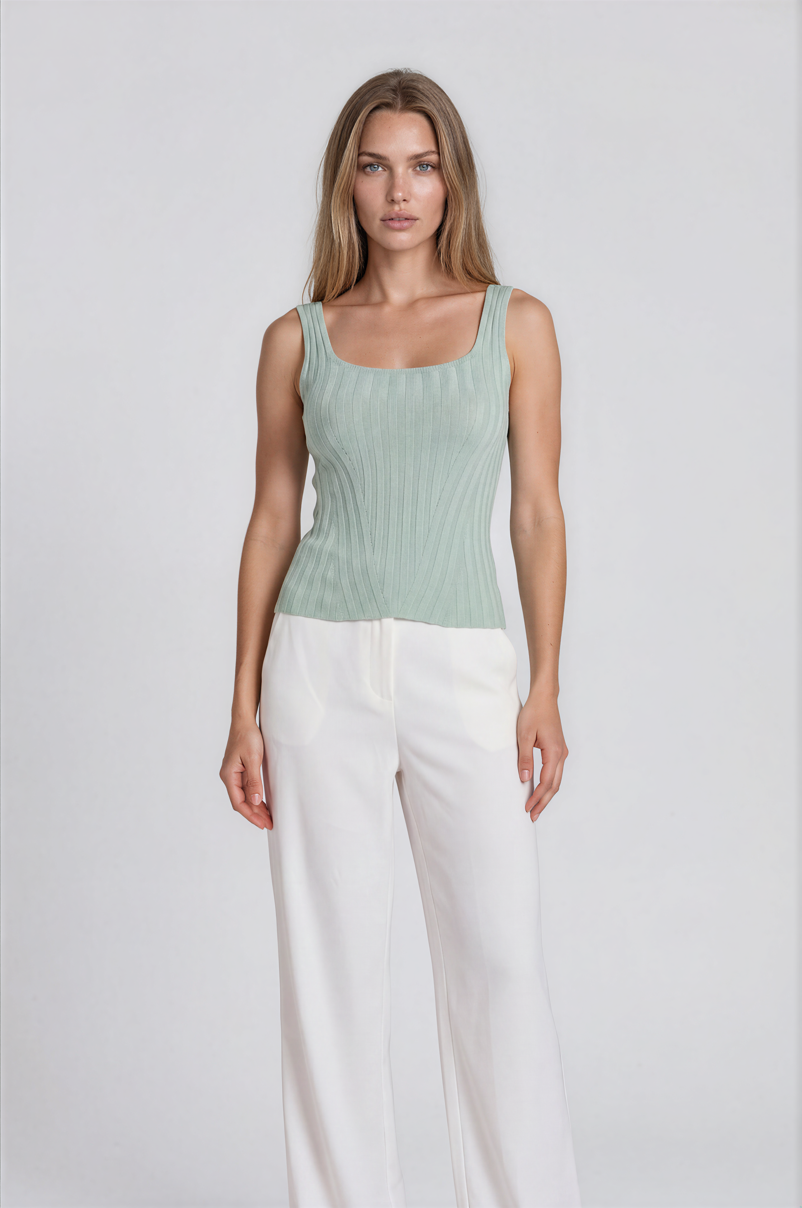 Woman wearing a light green sleeveless top and white pants on a plain background