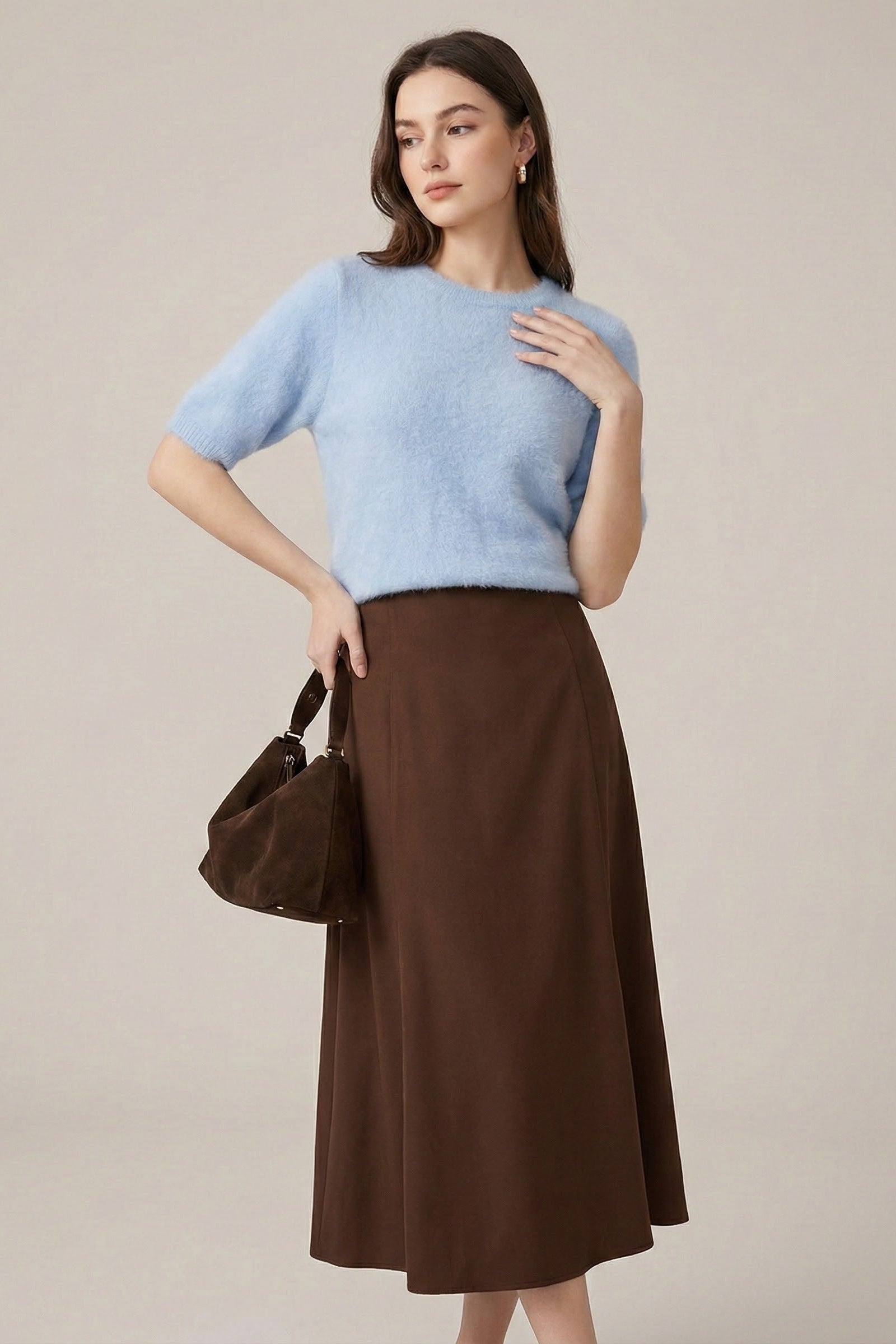 Woman wearing a light blue sweater and brown skirt holding a brown handbag against a beige background