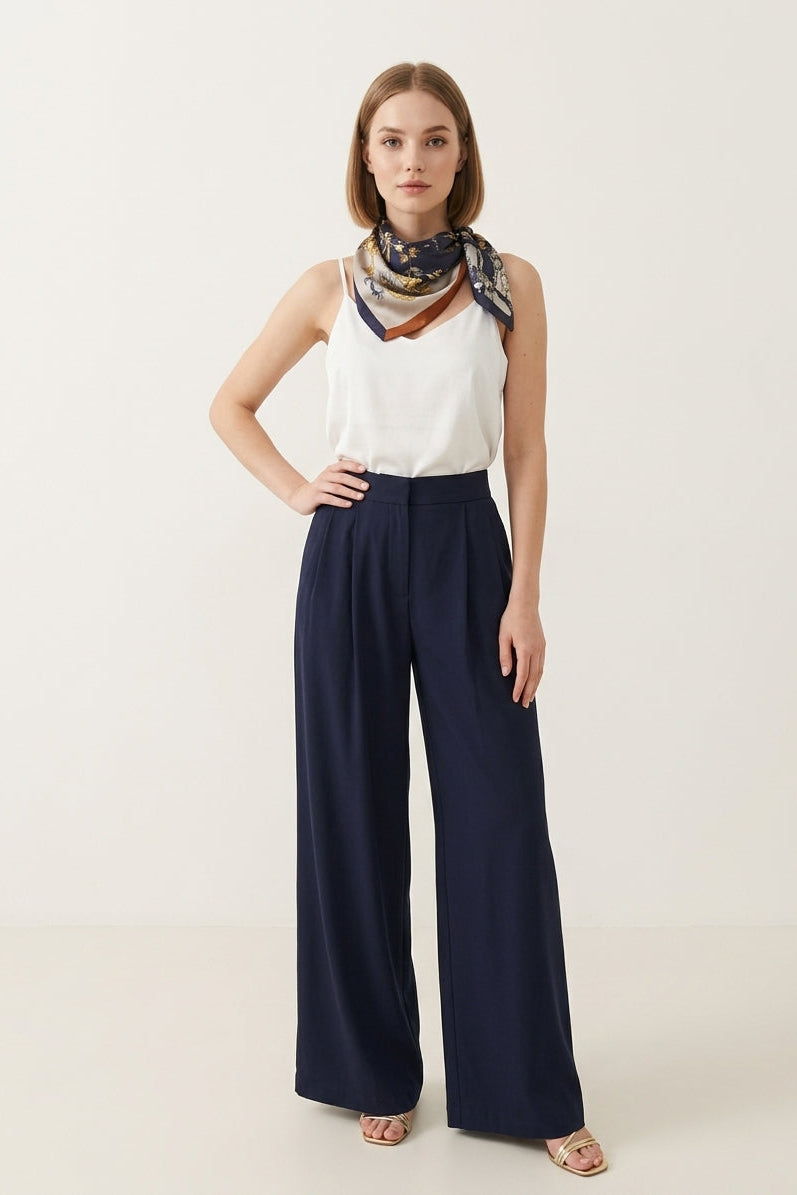 Woman wearing a white sleeveless top and navy wide-leg pants with a patterned scarf on a light background