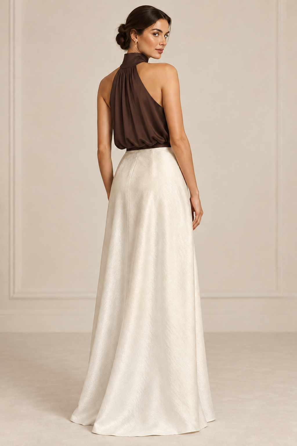 A woman modeling a floor-length formal dress from the back, featuring a dark brown draped halter-style bodice and a flowing ivory or off-white skirt, set against a neutral cream paneled studio background.