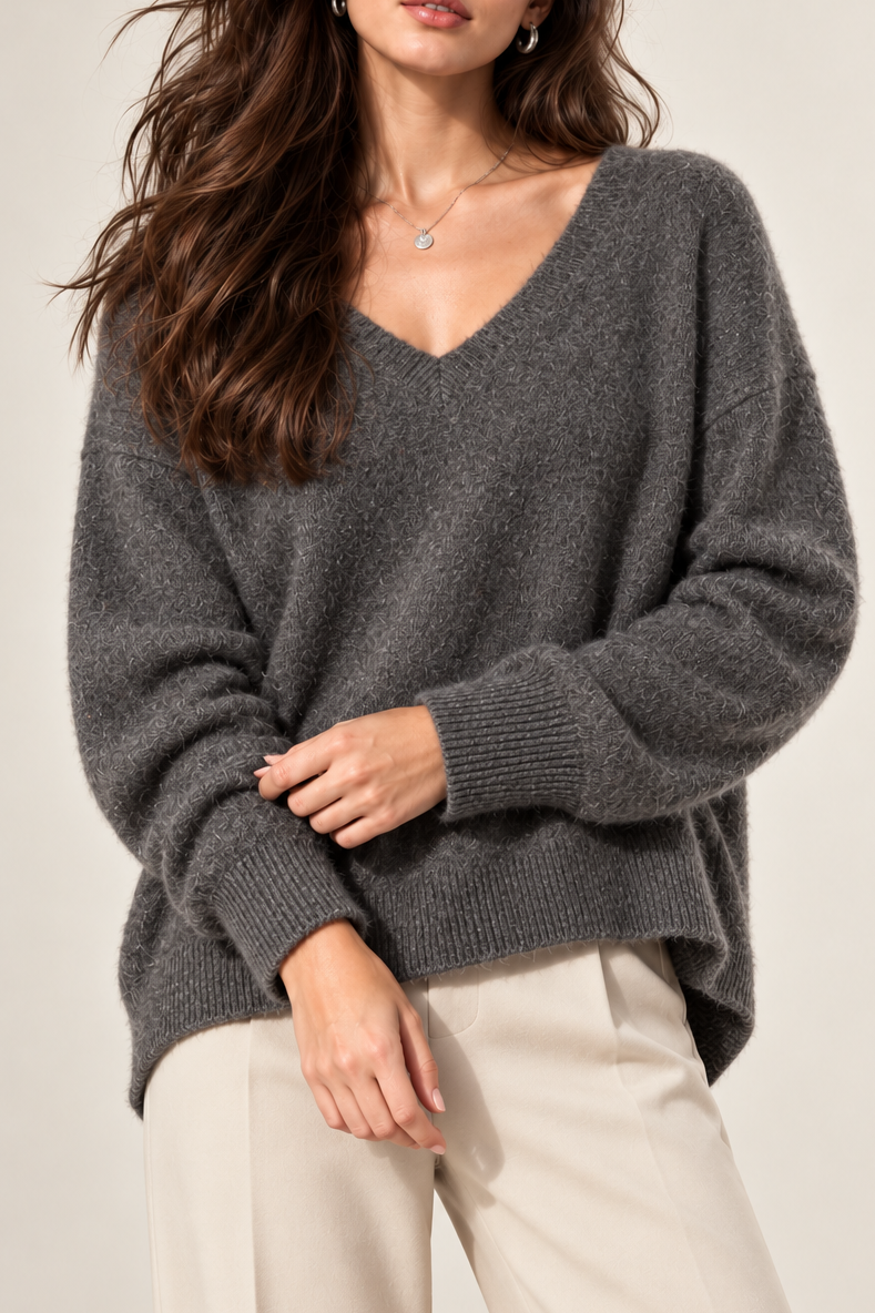 A woman modeling a dark gray heathered knit sweater with a wide V-neck and long sleeves, paired with light beige trousers, against a plain light beige studio background.
