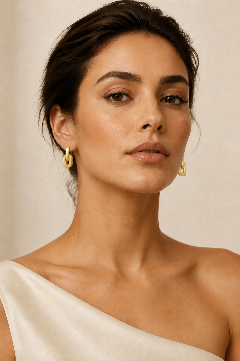 A close-up portrait of a woman wearing small gold hoop earrings against a soft beige background.