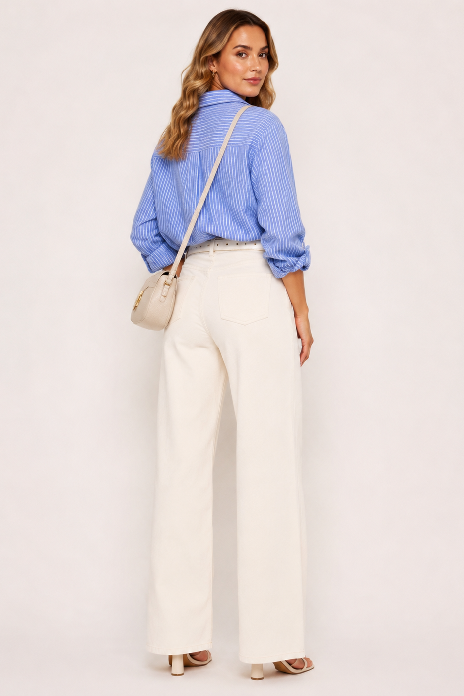 A woman is shown from the back wearing a light blue and white striped button-down shirt, high-waisted off-white wide-leg trousers, and carrying a small beige shoulder bag against a plain light background.