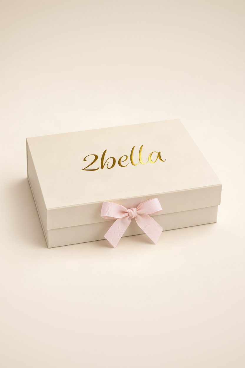 A cream-colored rectangular gift box with the name '2bella' printed in elegant gold foil script on the lid, tied with a light pink satin ribbon bow at the front, set against a solid cream background.