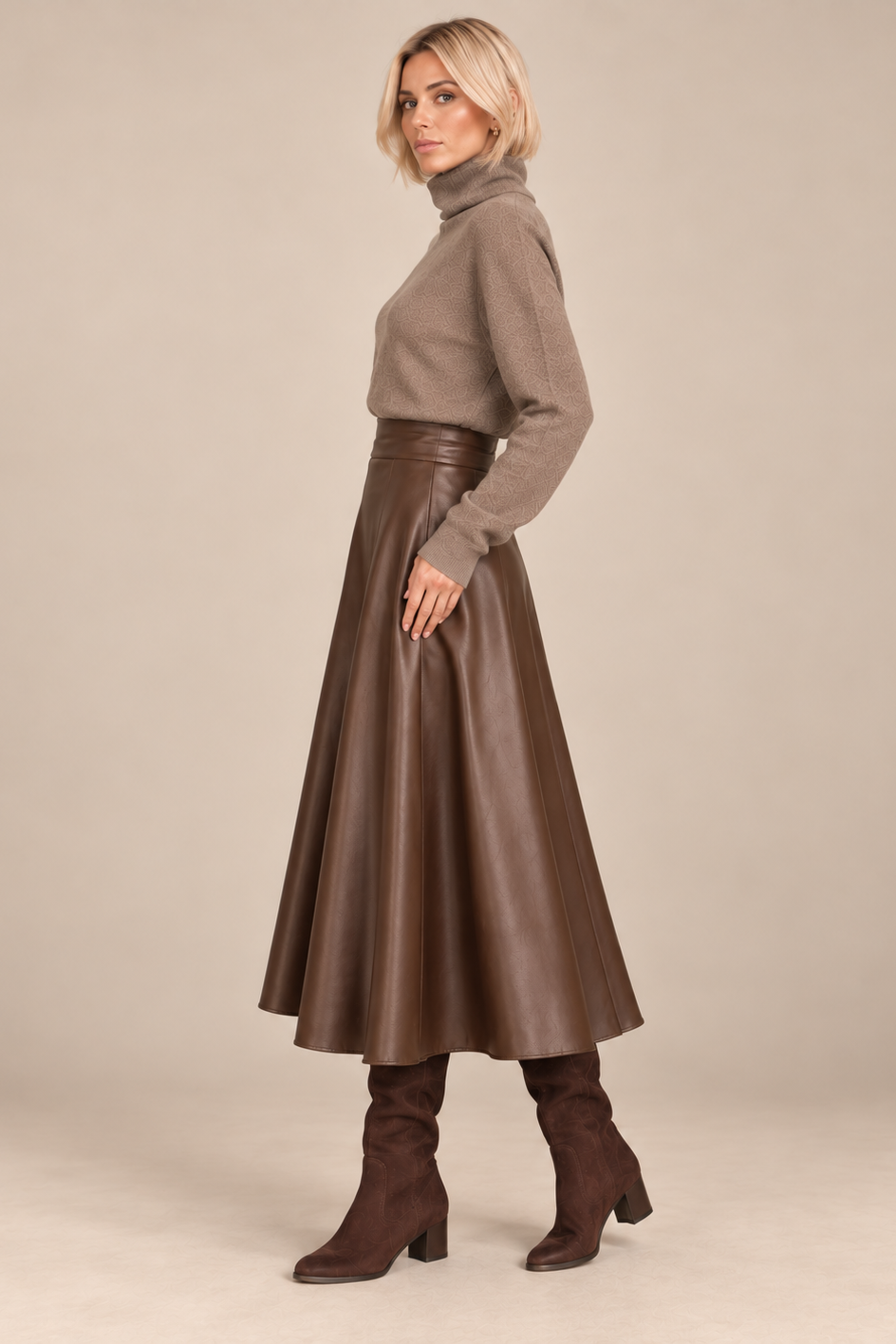 A woman stands in profile wearing a taupe turtleneck sweater, a brown faux-leather midi skirt with a matching belt, and brown heeled boots against a plain beige studio background.
