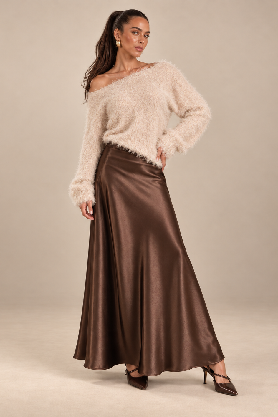 A woman models a two-piece outfit consisting of a fuzzy off-the-shoulder cream sweater and a high-waisted, floor-length brown satin skirt, set against a plain beige studio background.