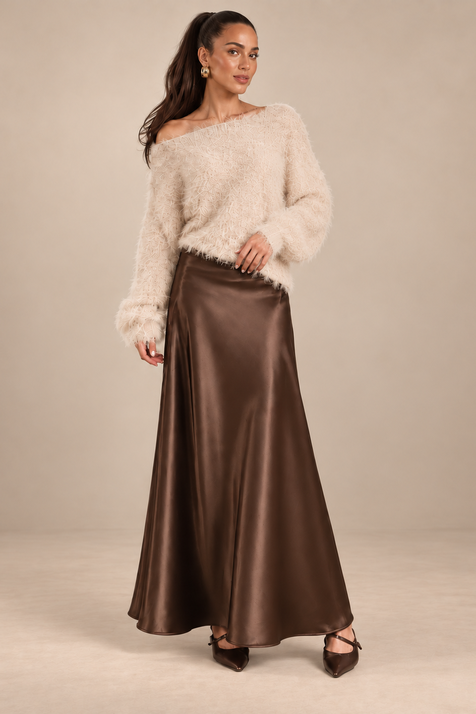 A woman modeling a cream fuzzy off-the-shoulder sweater and a brown satin maxi skirt against a neutral beige studio background.