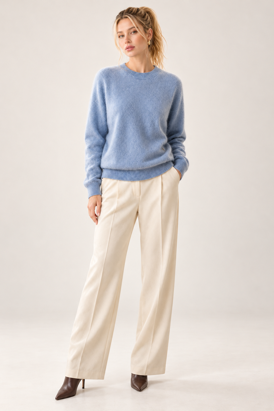 A woman modeling a light blue crewneck sweater and cream wide-leg trousers with brown pointed-toe heels, standing against a plain light gray studio background.