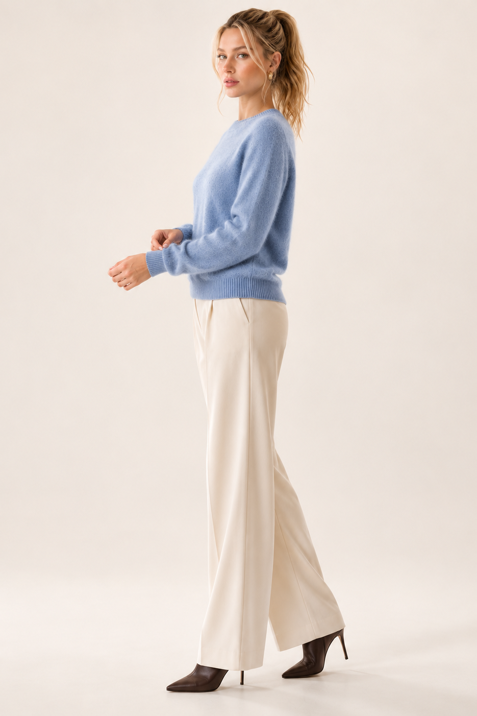 A woman stands in profile wearing a light blue knit sweater, wide-leg cream trousers, and dark brown pointed-toe heels against a plain warm off-white studio background.