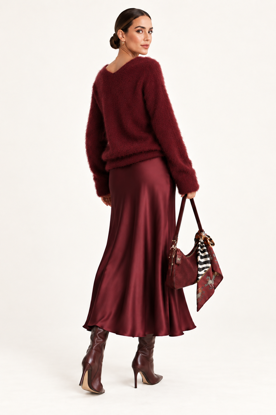 A woman stands in a studio wearing a fuzzy burgundy sweater, a satin-like burgundy midi skirt, and burgundy heeled boots, while holding a patterned handbag against a plain light background.