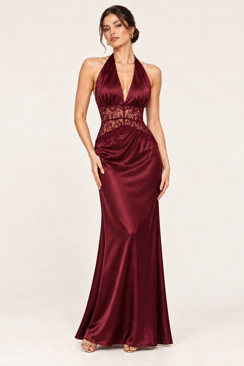 A woman modeling a floor-length halter gown in deep burgundy satin with a lace waist panel, photographed against a plain light studio background.