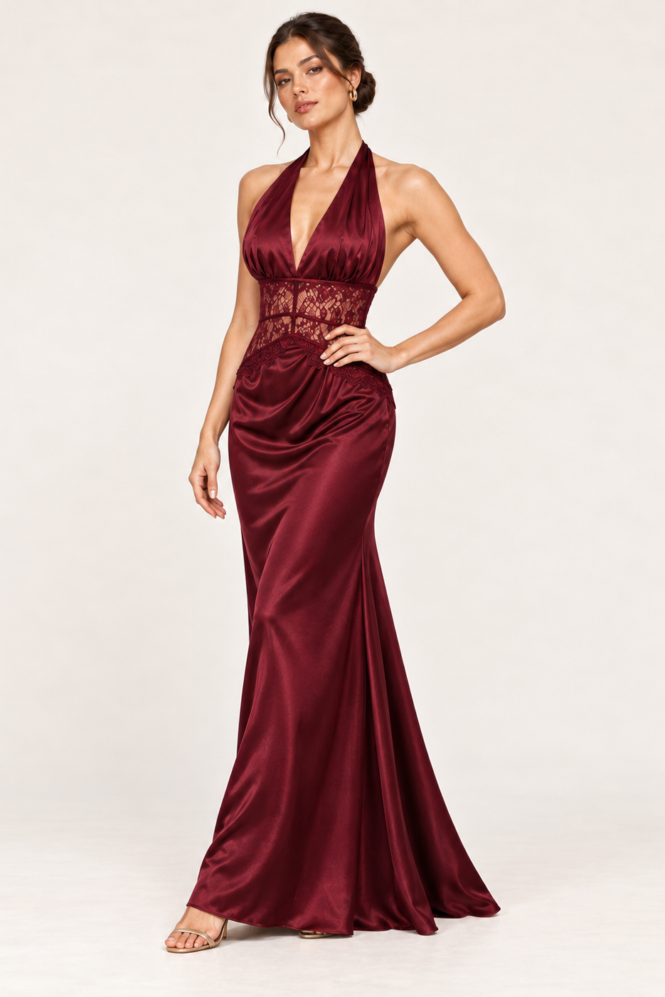 A woman models a floor-length halter gown in deep burgundy satin with a lace-embellished waistband, photographed against a clean off-white studio background.