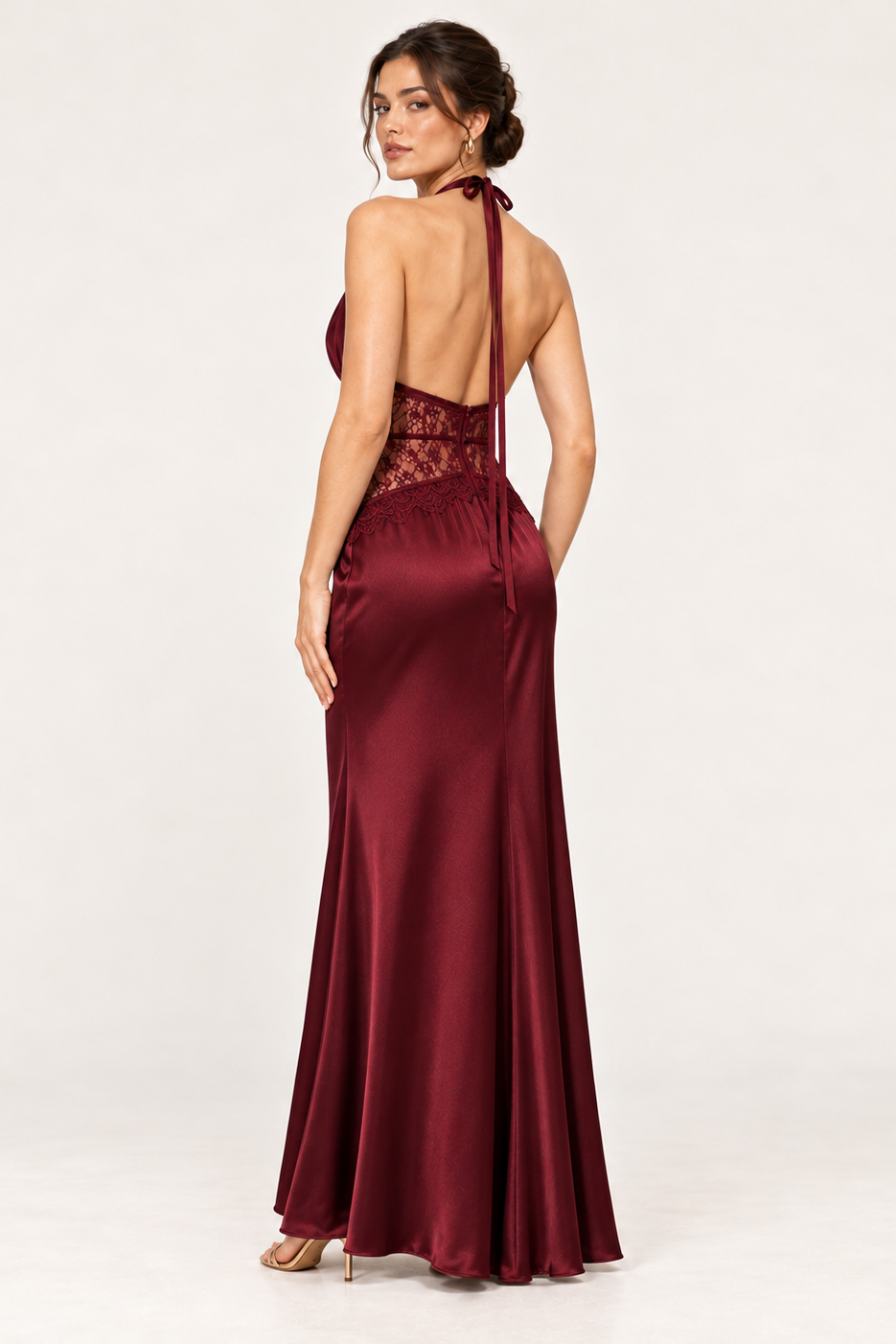 A woman modeling a floor-length burgundy satin evening gown from the back, featuring a lace back bodice, thin tie straps, and a flared skirt, set against a plain white studio background.