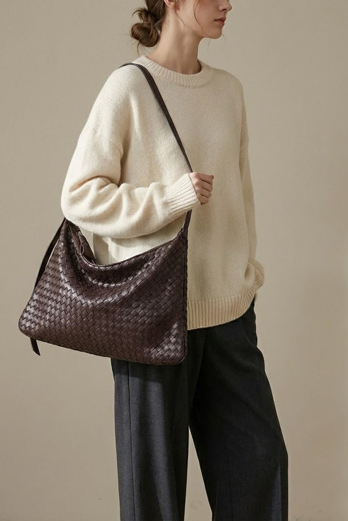 Person wearing a beige sweater and holding a woven brown bag against a plain background
