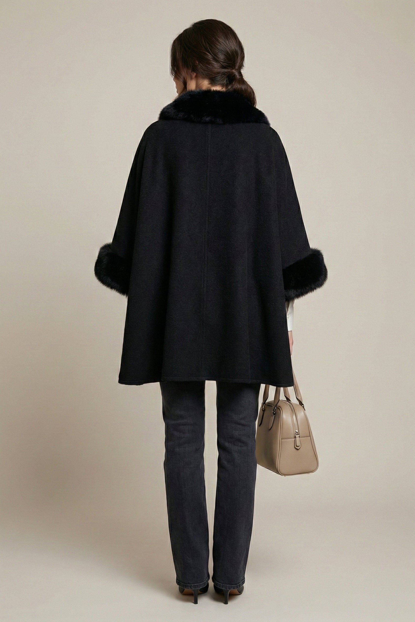 Person wearing a black coat with fur trim, holding a beige handbag on a beige background