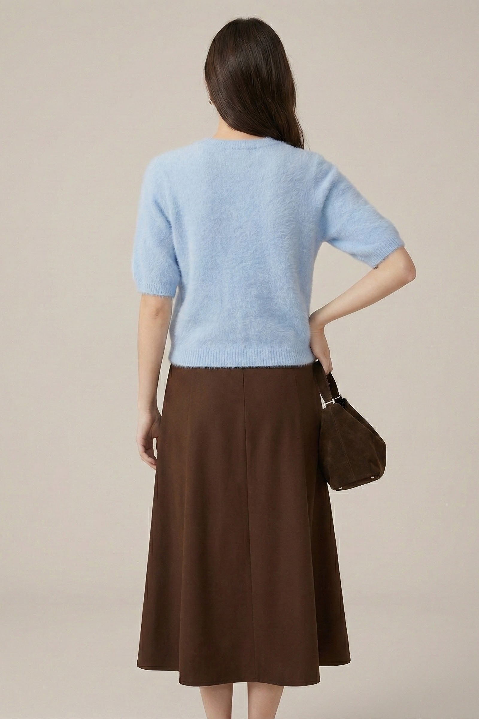 Woman wearing a light blue sweater and brown skirt with a handbag on a neutral background